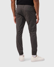 A person wearing Psycho Bunny MENS MADISON COMMUTER PANT - B6P395Z1WB in dark gray, along with white sneakers, is standing with their back to the camera. The pants feature an elastic waistband and cuffs, as well as a small zipper pocket on the back left side. They are standing against a plain light gray background adorned with subtle Bunny detailing.