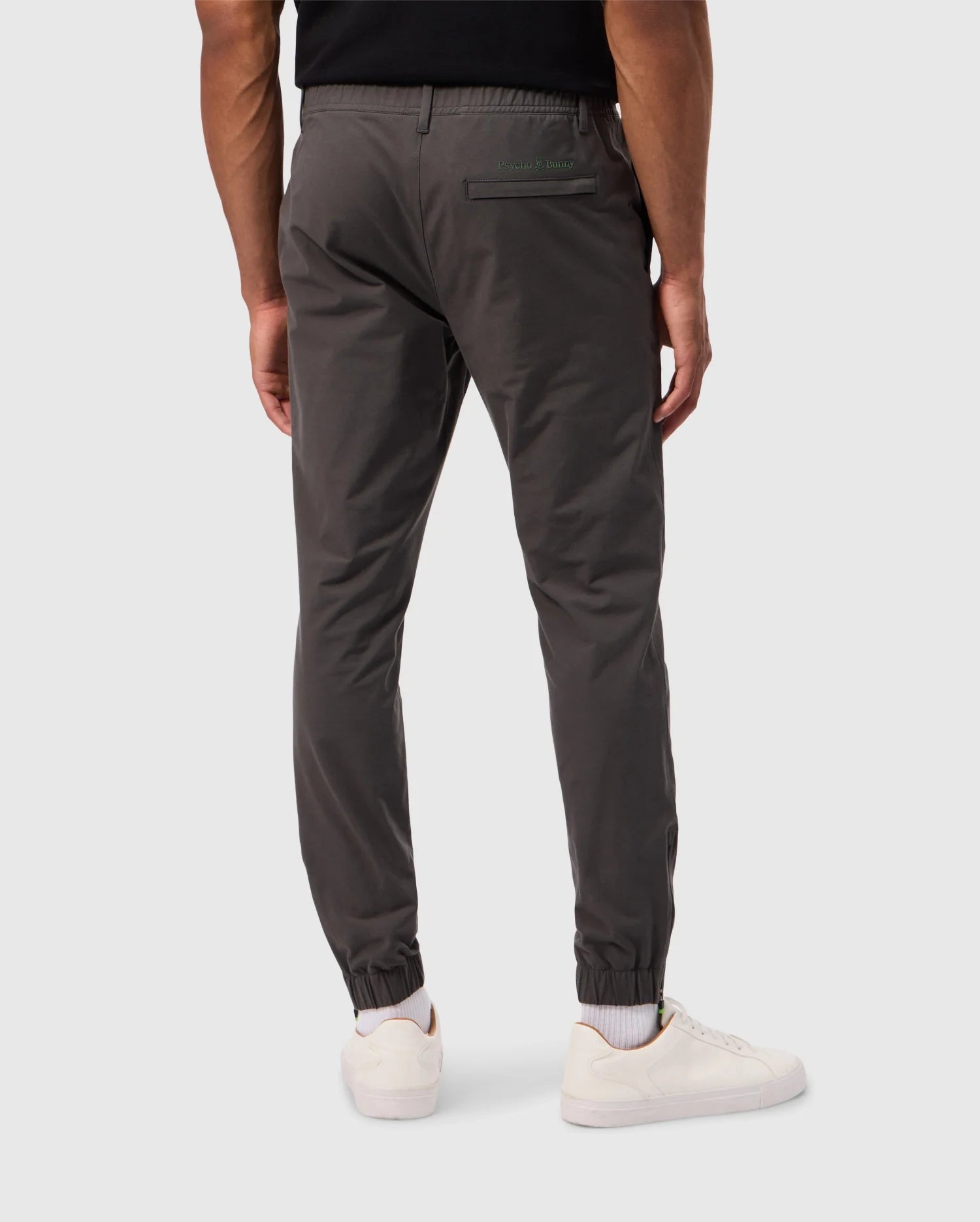 A person wearing Psycho Bunny MENS MADISON COMMUTER PANT - B6P395Z1WB in dark gray, along with white sneakers, is standing with their back to the camera. The pants feature an elastic waistband and cuffs, as well as a small zipper pocket on the back left side. They are standing against a plain light gray background adorned with subtle Bunny detailing.