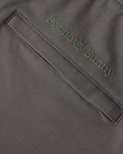 Close-up of a garment pocket with an embroidered "Psycho Bunny" logo above it, featuring the brand name in green letters and a bunny skull and crossbones design. The dark and smooth fabric suggests it could be from the MENS MADISON COMMUTER PANT - B6P395Z1WB, adorned with subtle Bunny detailing.