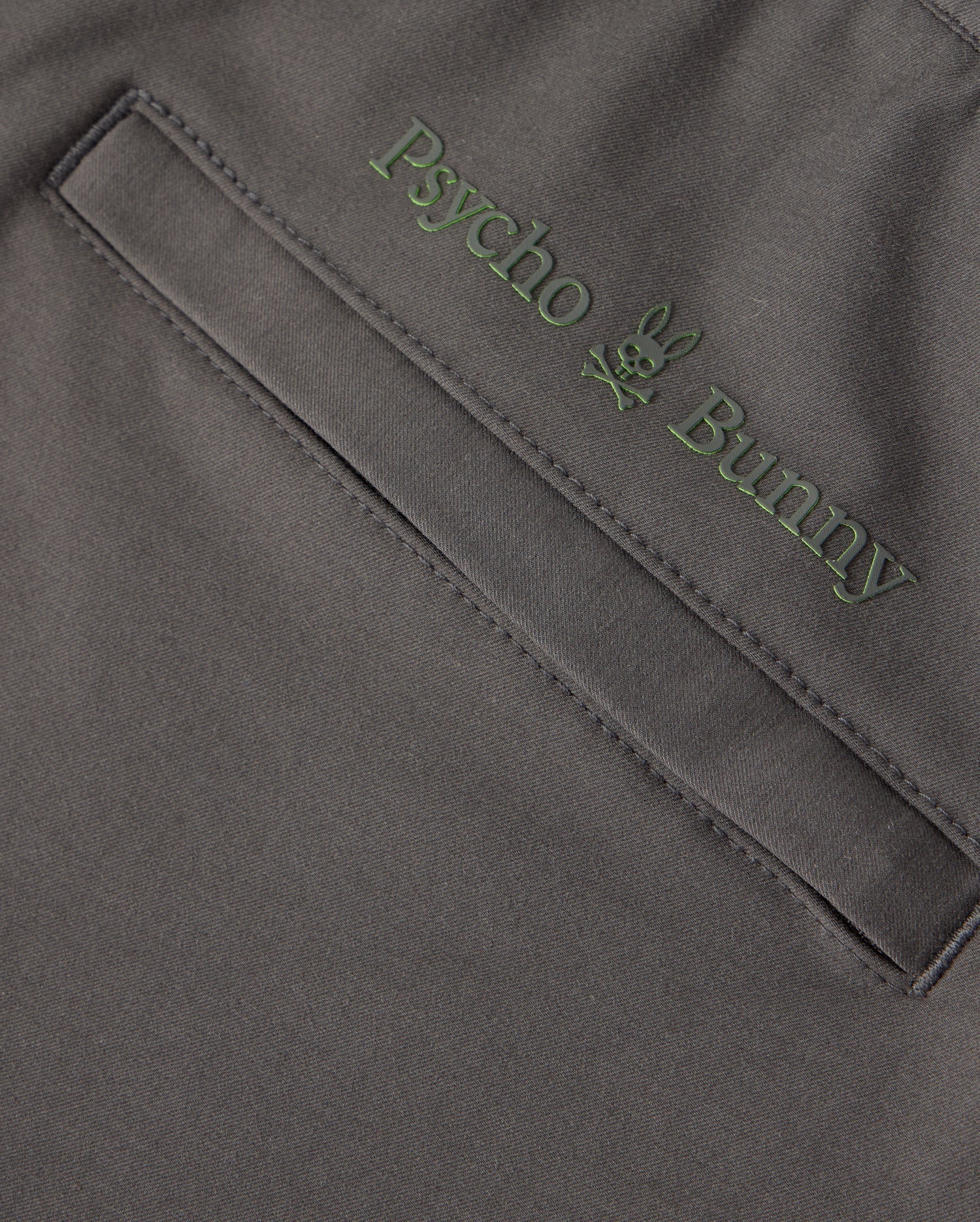 Close-up of a garment pocket with an embroidered "Psycho Bunny" logo above it, featuring the brand name in green letters and a bunny skull and crossbones design. The dark and smooth fabric suggests it could be from the MENS MADISON COMMUTER PANT - B6P395Z1WB, adorned with subtle Bunny detailing.