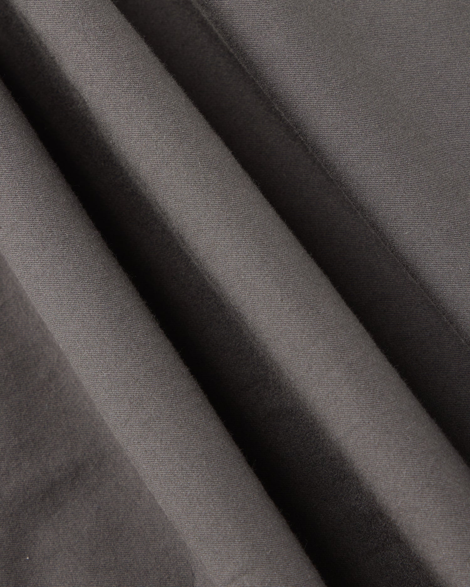 Close-up of smooth, dark grey fabric with soft wrinkles and folds, showcasing the fine texture and matte finish. This material, part of the MENS MADISON COMMUTER PANT - B6P395Z1WB by Psycho Bunny, creates a sense of depth and shadow throughout the image. Look closely to find subtle bunny detailing that adds a unique touch.