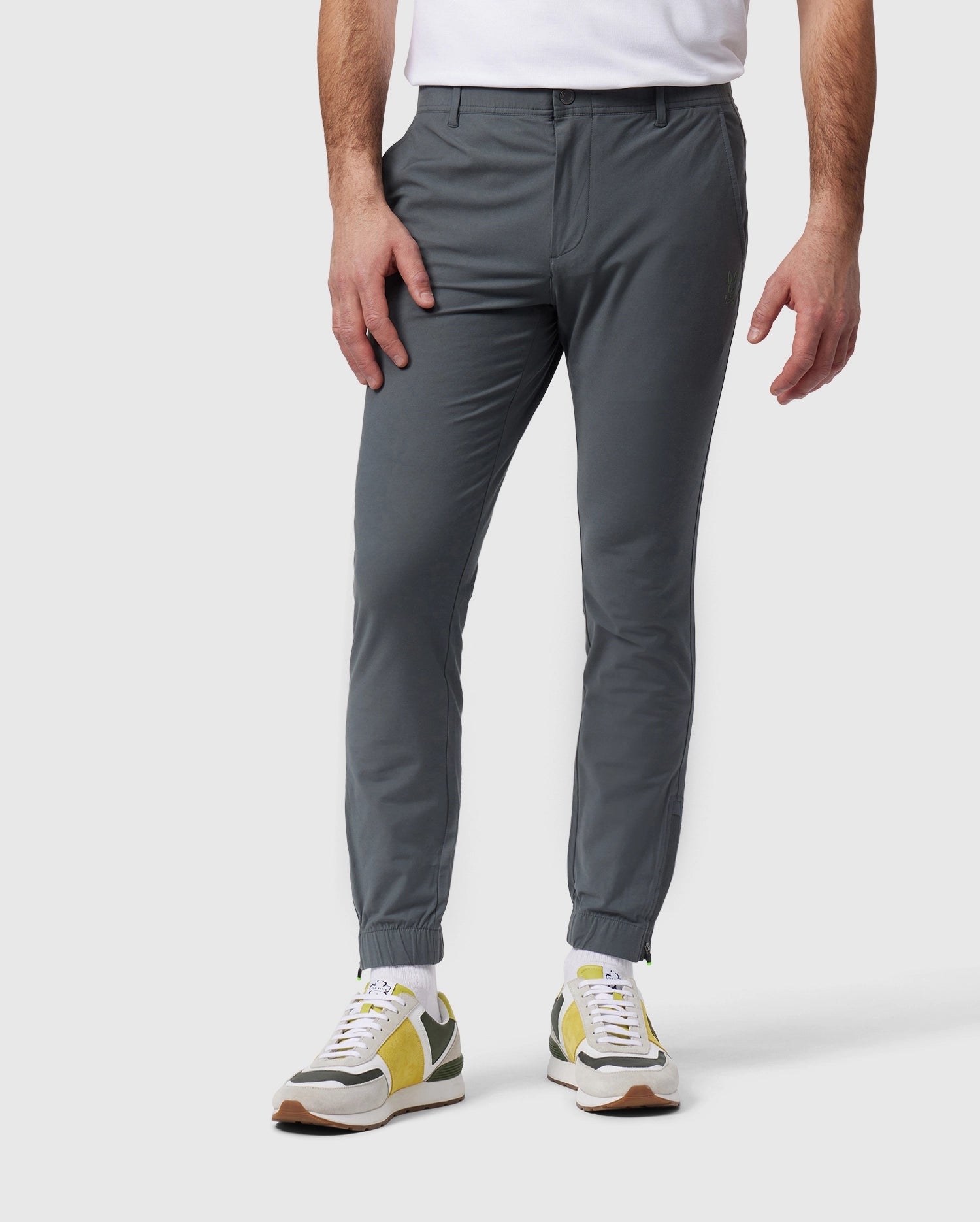 In a plain gray setting, a person wears Psycho Bunny's MENS MADISON COMMUTER PANT (B6P395Z1WB) made of gray stretch woven fabric and featuring an elasticized waistband. The look is completed with green and white sneakers, hands at their sides, and a partially visible white shirt.