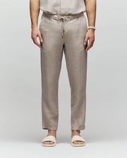 A model, visible from the shoulders down, wears Psycho Bunny's MENS FATE LINEN DRAWSTRING PANT (B6P471C200) with a matching embroidered shirt and cream slide sandals, standing against a plain light gray background.