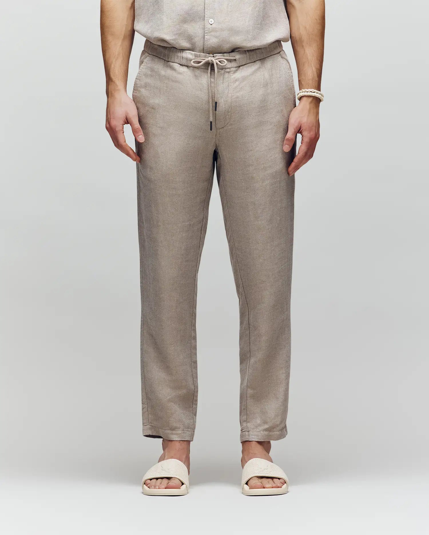 A model, visible from the shoulders down, wears Psycho Bunny's MENS FATE LINEN DRAWSTRING PANT (B6P471C200) with a matching embroidered shirt and cream slide sandals, standing against a plain light gray background.