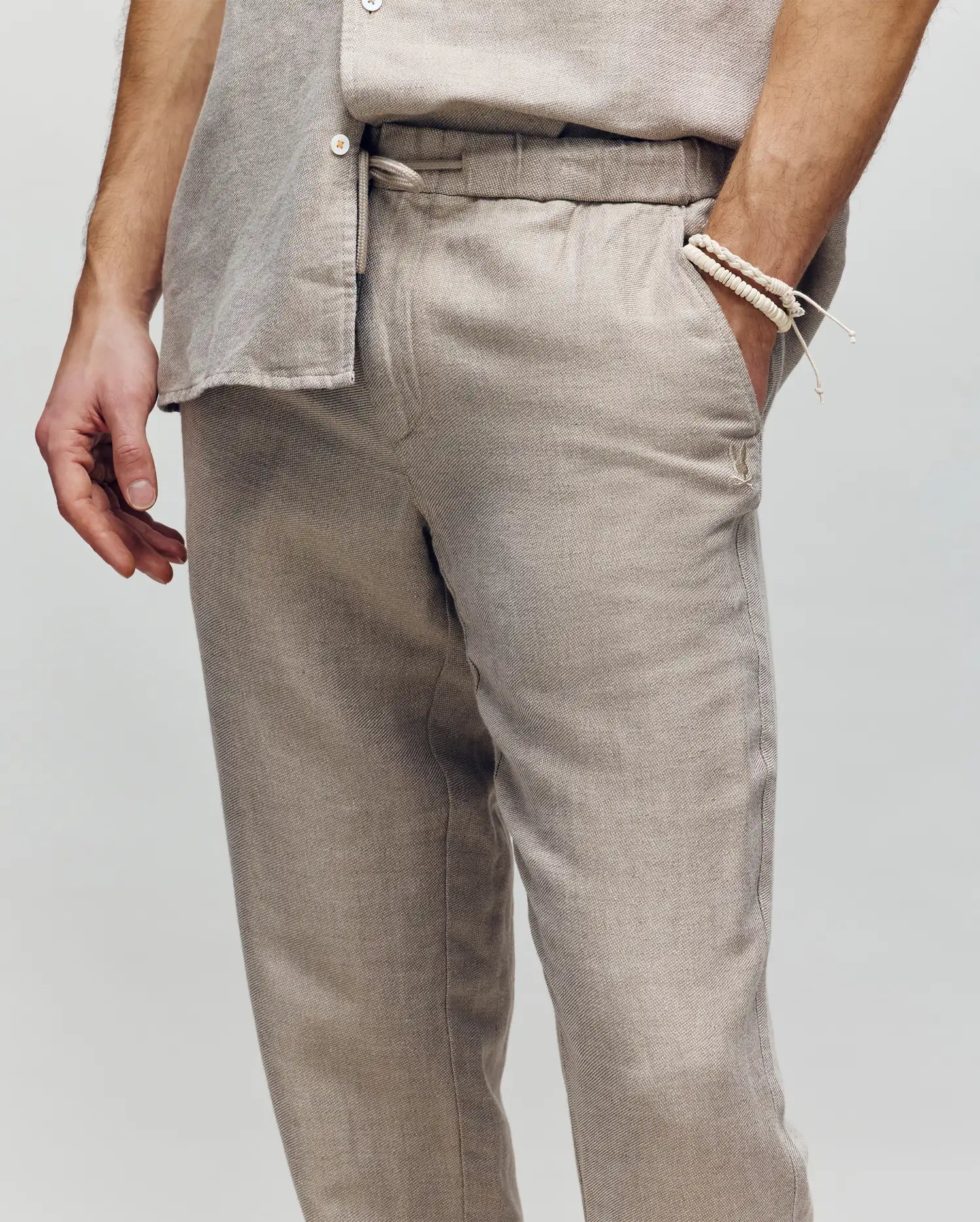 A person wears Psycho Bunny's MENS FATE LINEN DRAWSTRING PANT (B6P471C200) in light beige with a matching shirt, styled simply with a beaded bracelet and embroidered logo, standing against a plain neutral background.