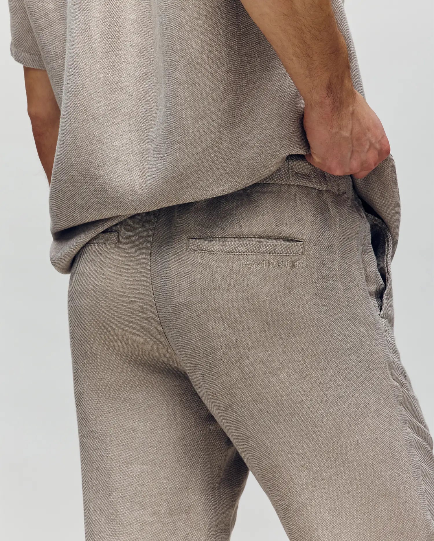 A person models the Psycho Bunny MENS FATE LINEN DRAWSTRING PANT (B6P471C200) in light beige, with a matching shirt. One hand rests in their pocket, highlighting the back pocket and side. The plain, light background keeps focus on the pants.