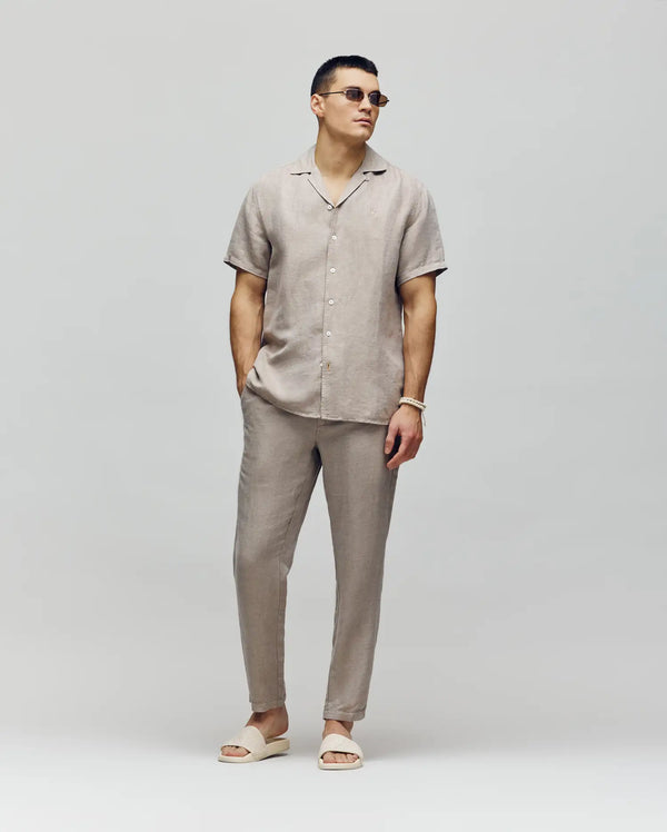 A man wearing Psycho Bunny's MENS FATE LINEN DRAWSTRING PANT (B6P471C200) in beige, a matching linen blend short-sleeve shirt, and beige slide sandals stands against a plain light background with his hands in his pockets.