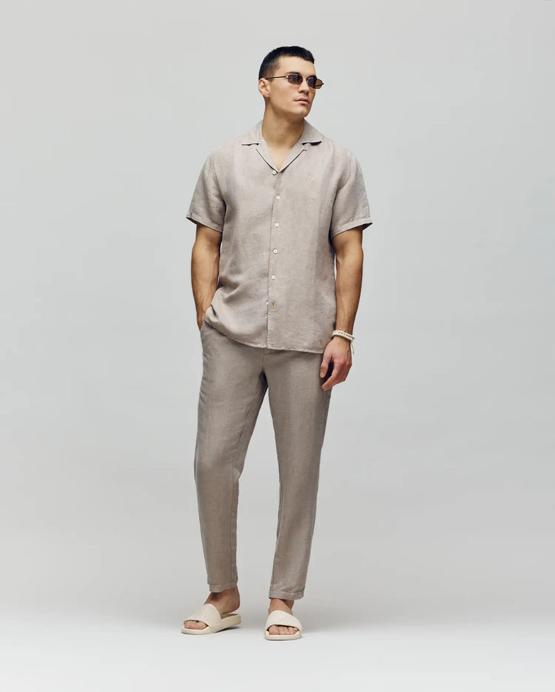 A man wearing Psycho Bunny's MENS FATE LINEN DRAWSTRING PANT (B6P471C200) in beige, a matching linen blend short-sleeve shirt, and beige slide sandals stands against a plain light background with his hands in his pockets.