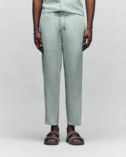 A person models only the lower half in Psycho Bunny's MENS FATE LINEN DRAWSTRING PANT - B6P471C200, light green and made from a cooling premium linen blend, paired with olive green sandals against a plain background.