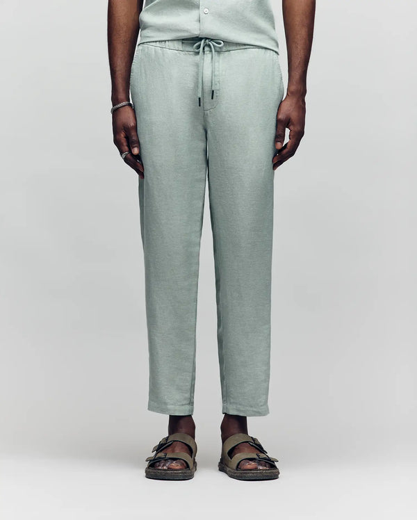 A person models only the lower half in Psycho Bunny's MENS FATE LINEN DRAWSTRING PANT - B6P471C200, light green and made from a cooling premium linen blend, paired with olive green sandals against a plain background.