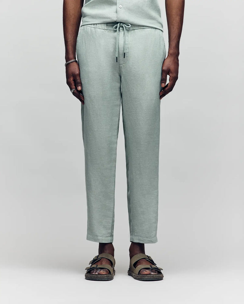 A person models only the lower half in Psycho Bunny's MENS FATE LINEN DRAWSTRING PANT - B6P471C200, light green and made from a cooling premium linen blend, paired with olive green sandals against a plain background.