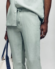 A person wears Psycho Bunny MENS FATE LINEN DRAWSTRING PANT - B6P471C200 in light green with a matching shirt, holding a white tote bag with navy handles in their left hand. Only the torso and upper legs are shown.