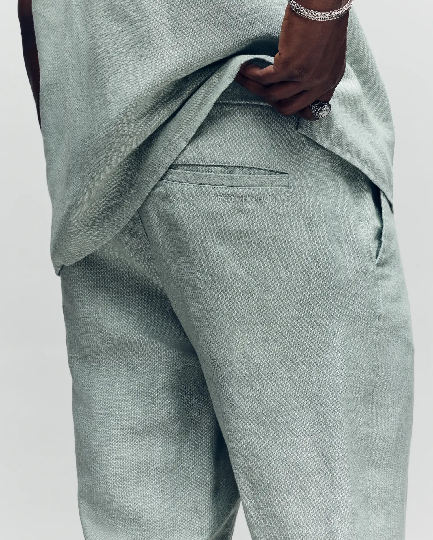 A man in light sage green Psycho Bunny MENS FATE LINEN DRAWSTRING PANT B6P471C200 lifts his matching shirt to show the back pocket with an embroidered logo. He's accessorized with a silver bracelet and ring.
