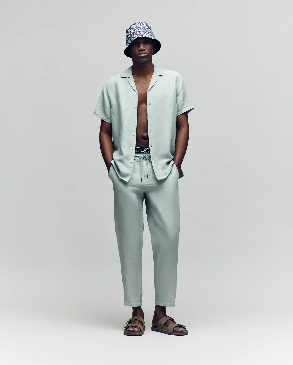 A man stands against a plain background, wearing Psycho Bunny’s MENS FATE LINEN DRAWSTRING PANT (B6P471C200) with a light gray linen-blend short-sleeve button-up shirt, a patterned bucket hat, sandals, and his hands in his pockets.