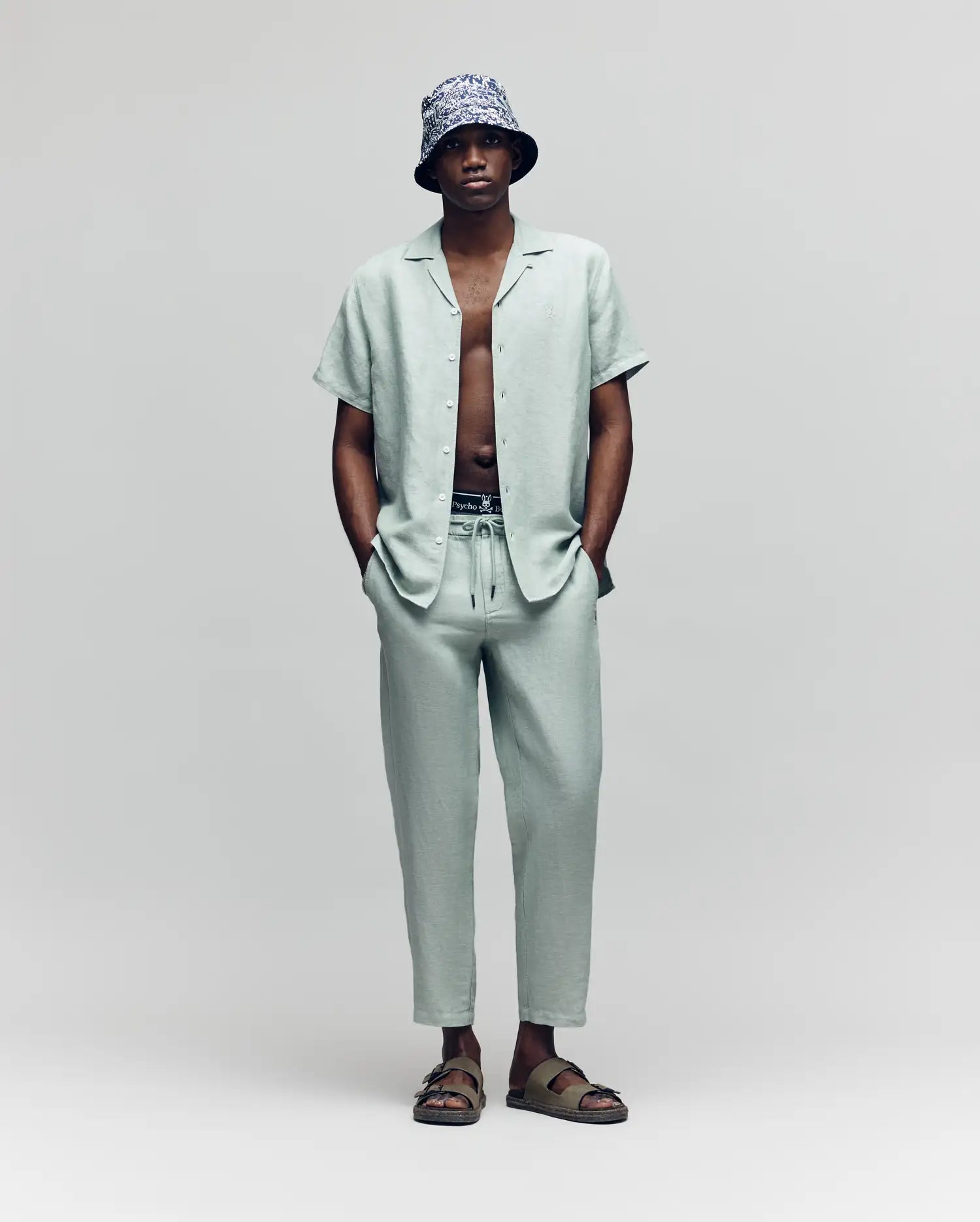 A man stands against a plain background, wearing Psycho Bunny’s MENS FATE LINEN DRAWSTRING PANT (B6P471C200) with a light gray linen-blend short-sleeve button-up shirt, a patterned bucket hat, sandals, and his hands in his pockets.