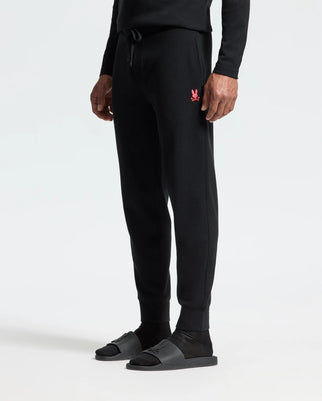 A person wearing the black Psycho Bunny MENS VAIL WAFFLE LOUNGE PANT - B6P673E200, featuring a small red logo on the left side, is paired with black socks and black slide sandals, standing against a plain white background.