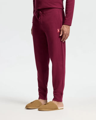 The person is wearing Psycho Bunny's maroon MENS VAL WAFFLE LOUNGE PANT, featuring a drawstring and small embroidered logo on the left thigh for textured comfort. They pair it with a matching long-sleeve top and tan slip-on shoes against a light gray backdrop.