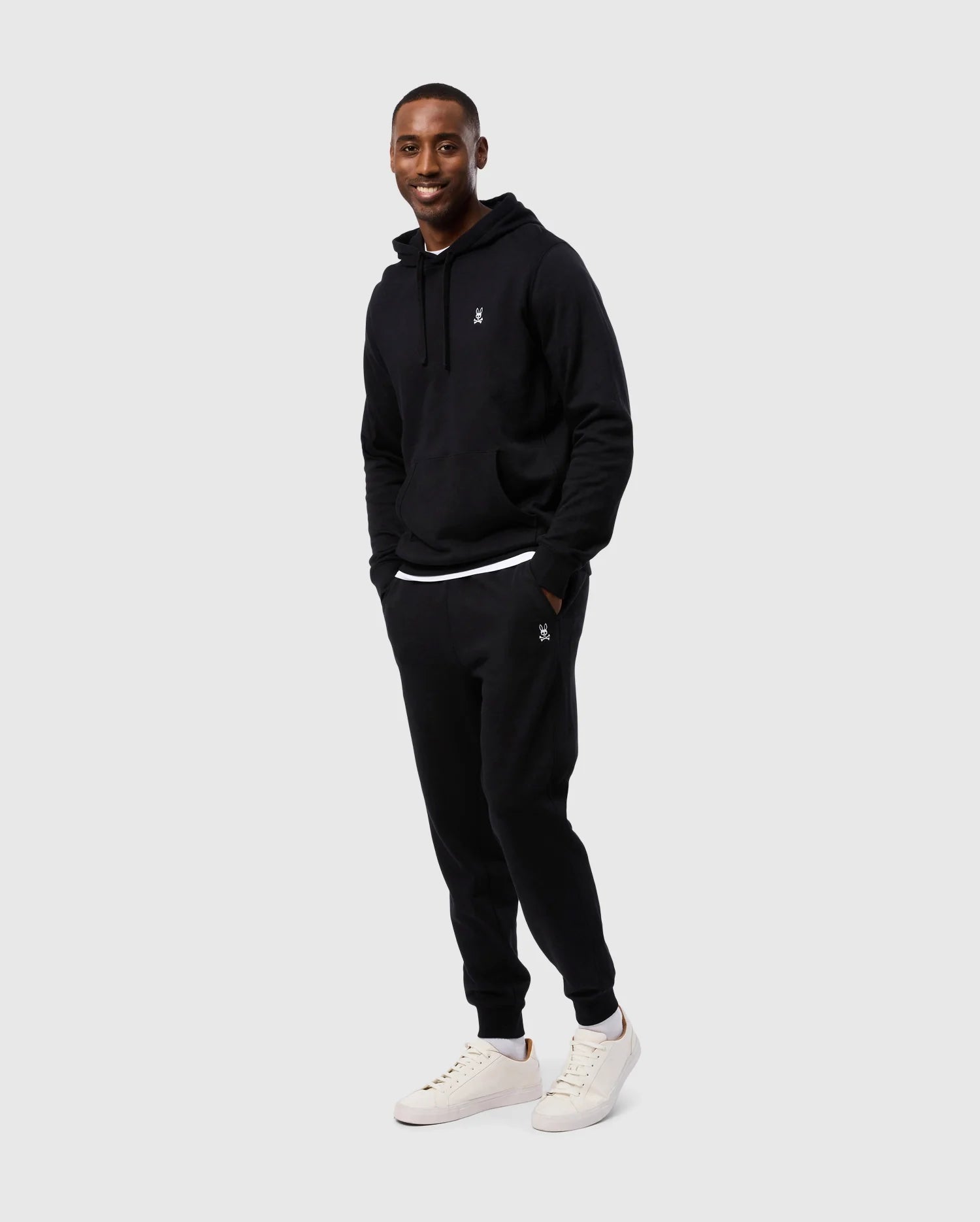 A person smiles while standing with hands in the pockets of a black hoodie and matching MENS LEVY ESSENTIAL FLEECE SWEATPANT (B6P721D200) by Psycho Bunny. They are wearing white sneakers. The background is plain and light-colored, perfectly showcasing their casual wear.