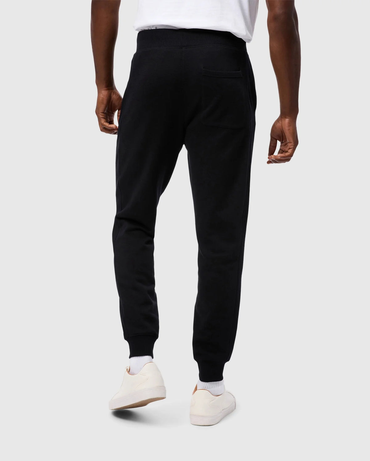 A person is standing facing away from the camera, wearing Psycho Bunny MENS LEVY ESSENTIAL FLEECE SWEATPANT in black (B6P721D200), white socks, and white sneakers. They are also wearing a white t-shirt. The background is a plain, light grey color.