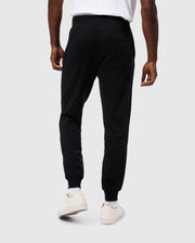 A person is standing facing away from the camera, wearing Psycho Bunny MENS LEVY ESSENTIAL FLEECE SWEATPANT in black (B6P721D200), white socks, and white sneakers. They are also wearing a white t-shirt. The background is a plain, light grey color.