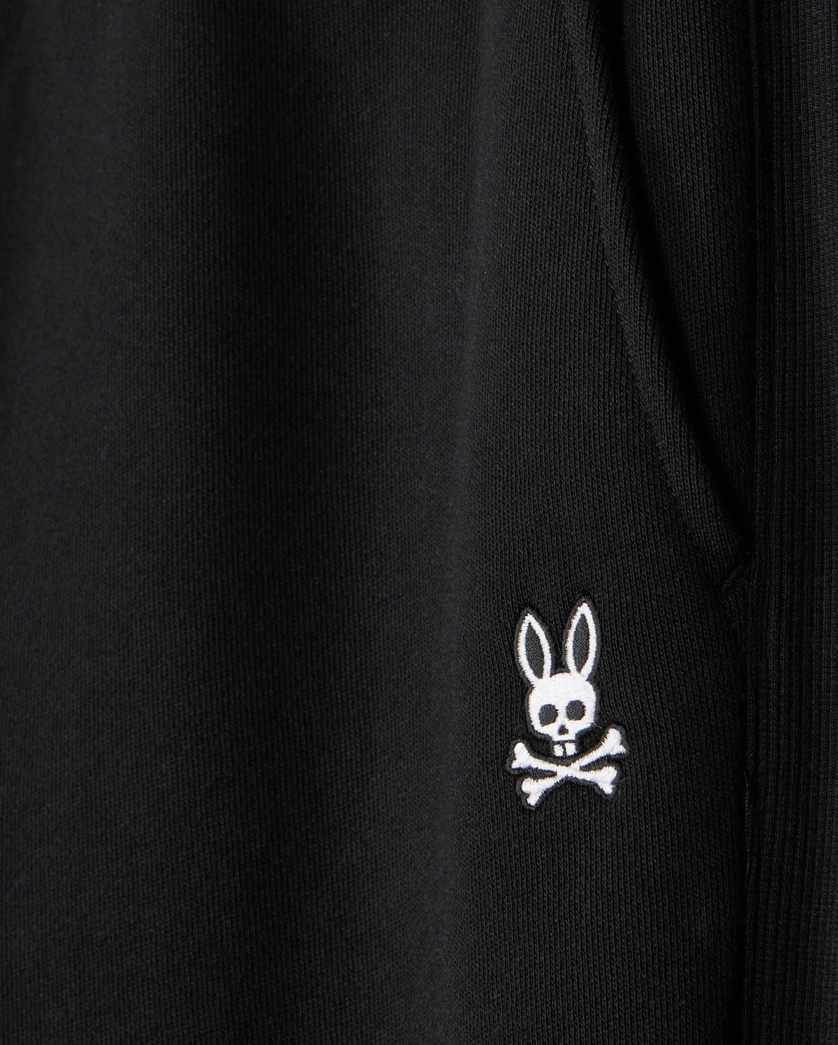Close-up of a black fabric on the Psycho Bunny MENS LEVY ESSENTIAL FLEECE SWEATPANT - B6P721D200, featuring an embroidered white emblem of a rabbit's head with crossbones underneath. The rabbit head has hollow eyes and a small skull in place of its nose, creating a spooky yet playful design perfect for casual wear.