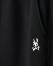 Close-up of a black fabric on the Psycho Bunny MENS LEVY ESSENTIAL FLEECE SWEATPANT - B6P721D200, featuring an embroidered white emblem of a rabbit's head with crossbones underneath. The rabbit head has hollow eyes and a small skull in place of its nose, creating a spooky yet playful design perfect for casual wear.