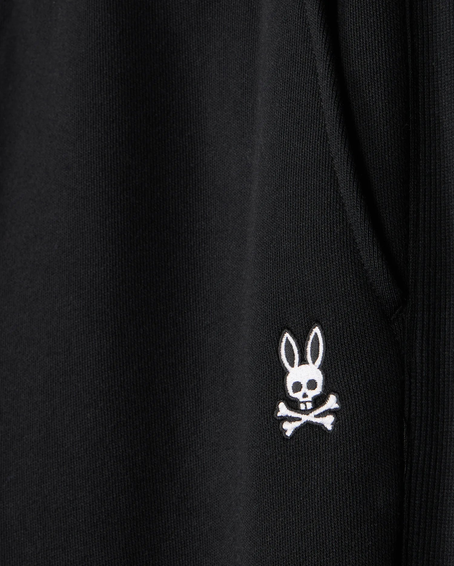 Close-up of a black fabric on the Psycho Bunny MENS LEVY ESSENTIAL FLEECE SWEATPANT - B6P721D200, featuring an embroidered white emblem of a rabbit's head with crossbones underneath. The rabbit head has hollow eyes and a small skull in place of its nose, creating a spooky yet playful design perfect for casual wear.