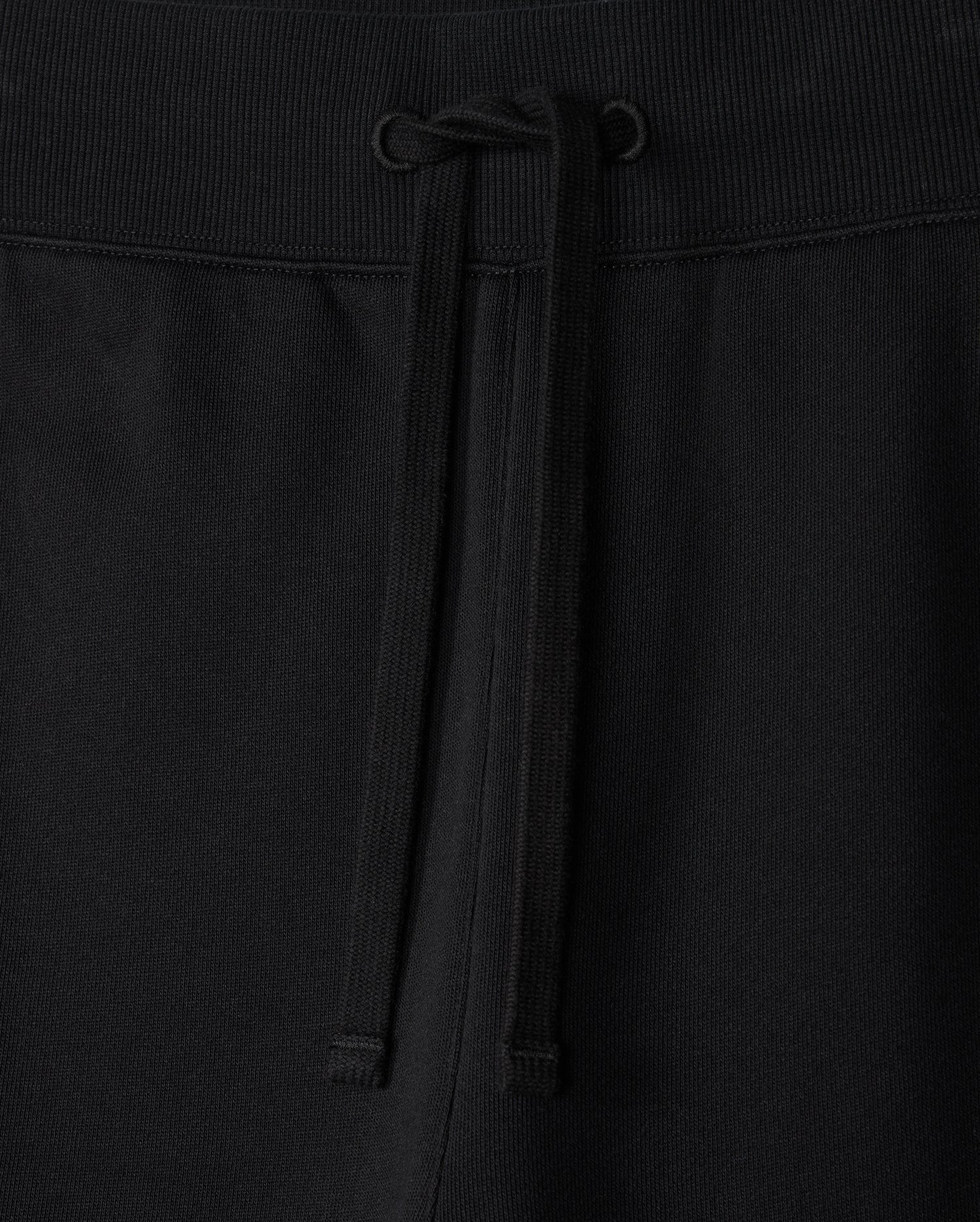 Close-up of the MENS LEVY ESSENTIAL FLEECE SWEATPANT - B6P721D200 by Psycho Bunny, showcasing black cotton sweatpants with an elastic waistband. The waistband features a matching black drawstring tied in a simple bow at the center. The texture of the fabric is visible, indicating comfort and durability, making them perfect for casual wear.