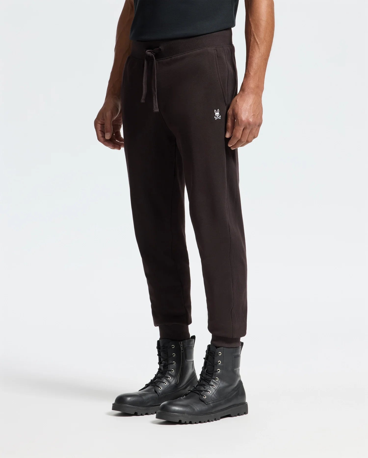 A person is wearing the Psycho Bunny Men's Levy Essential Fleece Sweatpant (B6P721D200) in black, made of 100% cotton with a small embroidered logo on the left side, paired with black lace-up combat boots. This casualwear look stands out against the plain white background.