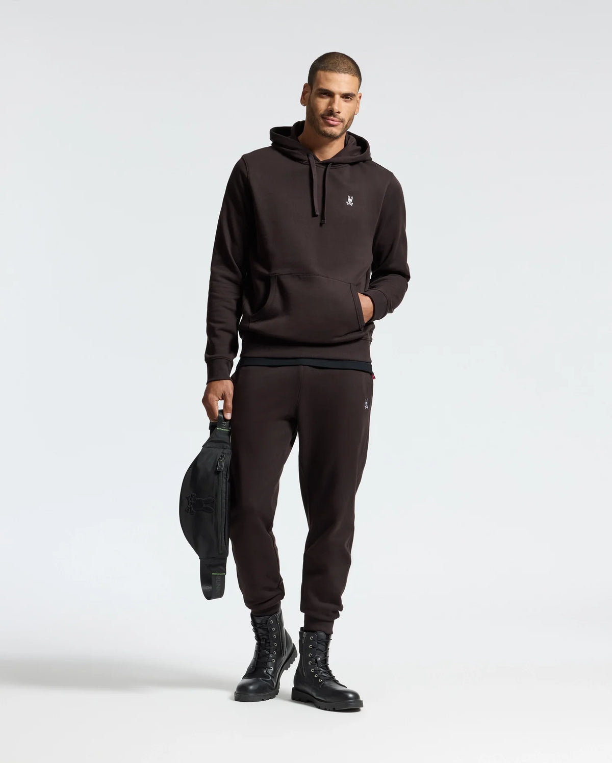 A man wearing a black hoodie and the MENS LEVY ESSENTIAL FLEECE SWEATPANT by Psycho Bunny stands against a plain background, holding a black bag. He is also sporting black lace-up boots and has one hand in his pocket, capturing the essence of casualwear.