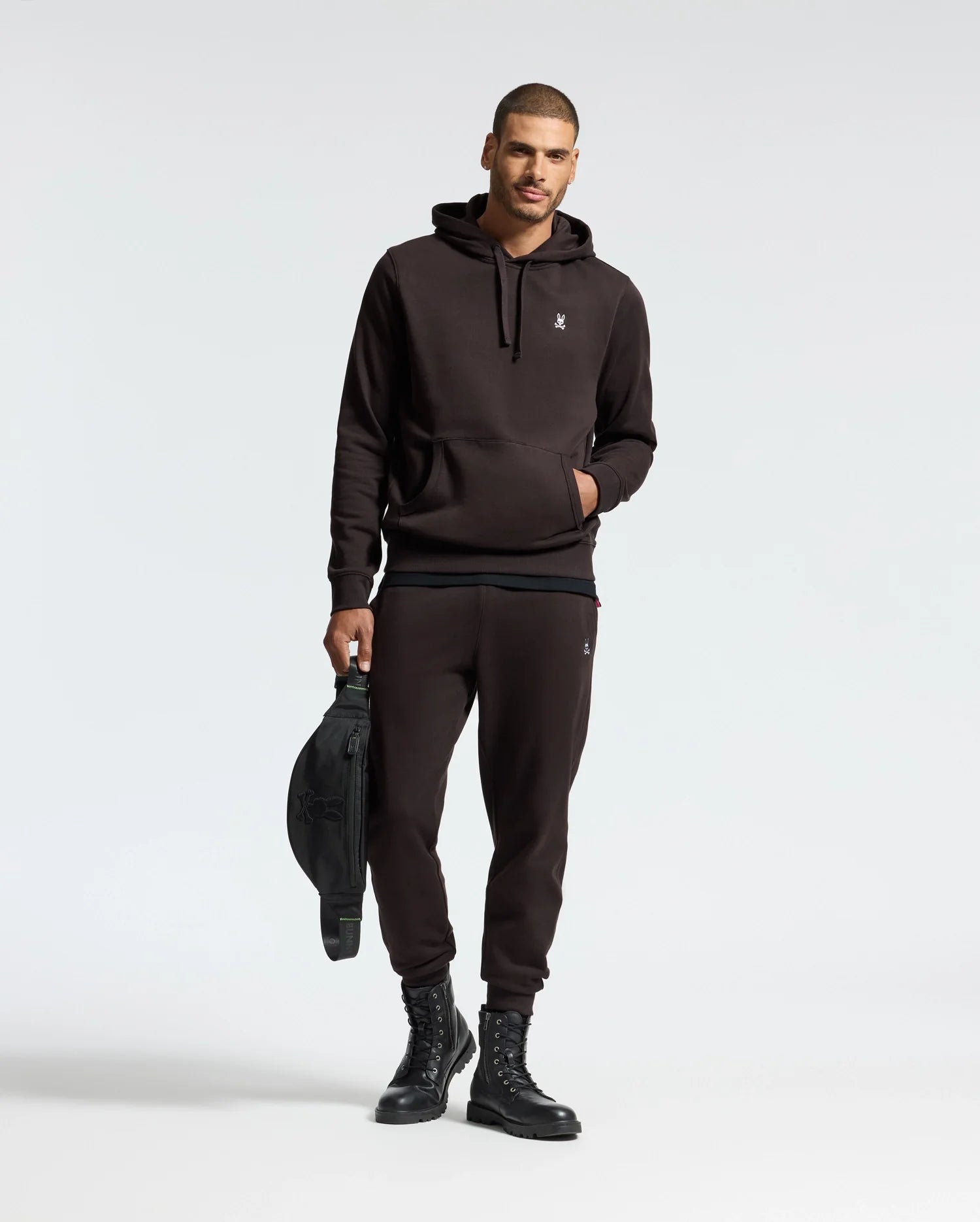 A man wearing a black hoodie and the MENS LEVY ESSENTIAL FLEECE SWEATPANT by Psycho Bunny stands against a plain background, holding a black bag. He is also sporting black lace-up boots and has one hand in his pocket, capturing the essence of casualwear.