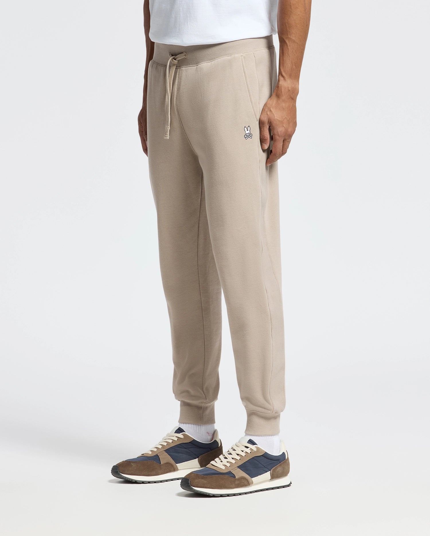 A person wearing Psycho Bunny's MENS LEVY ESSENTIAL FLEECE SWEATPANT - B6P721D200, featuring a chic embroidered logo near the pocket, paired with white socks and brown sneakers with black and white accents. The ensemble is completed with a white shirt as they stand against a plain background.
