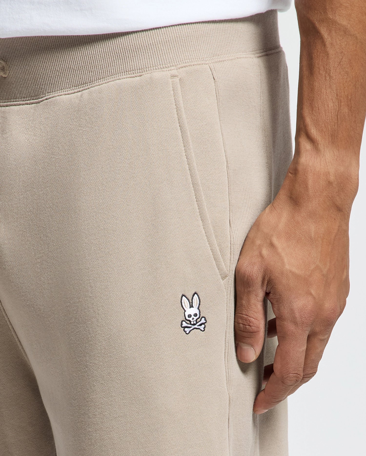 A close-up of someone wearing Psycho Bunny's MENS LEVY ESSENTIAL FLEECE SWEATPANT - B6P721D200, which are made from ultra-soft cotton fleece. The beige joggers display a small embroidered rabbit with crossed bones logo and have their hand nestled in the pocket. The look is finished off with a crisp white shirt.