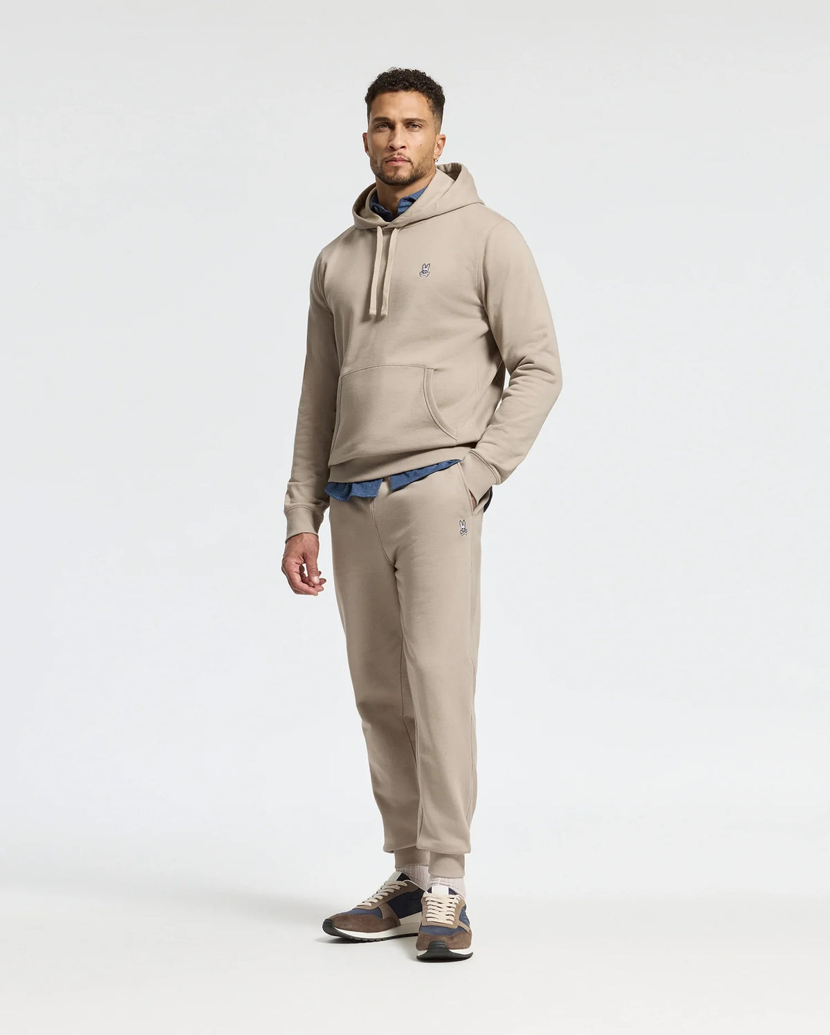A man stands against a plain white background wearing the MENS LEVY ESSENTIAL FLEECE SWEATPANT in beige, crafted from ultra-soft cotton fleece by Psycho Bunny. Beneath his hoodie, a blue shirt adds a pop of color to the ensemble, while sneakers complete this effortlessly stylish look.