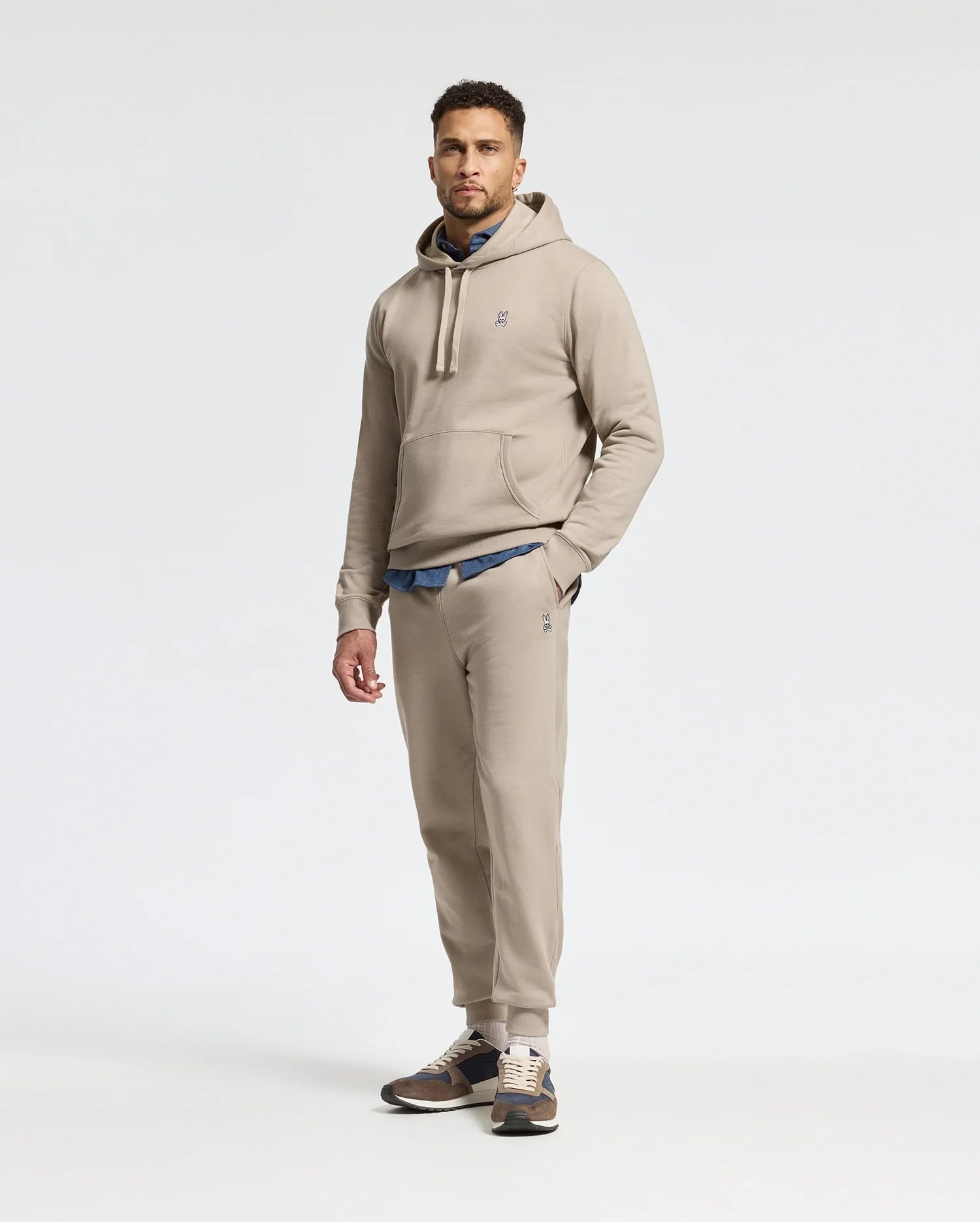 A man stands against a plain white background wearing the MENS LEVY ESSENTIAL FLEECE SWEATPANT in beige, crafted from ultra-soft cotton fleece by Psycho Bunny. Beneath his hoodie, a blue shirt adds a pop of color to the ensemble, while sneakers complete this effortlessly stylish look.