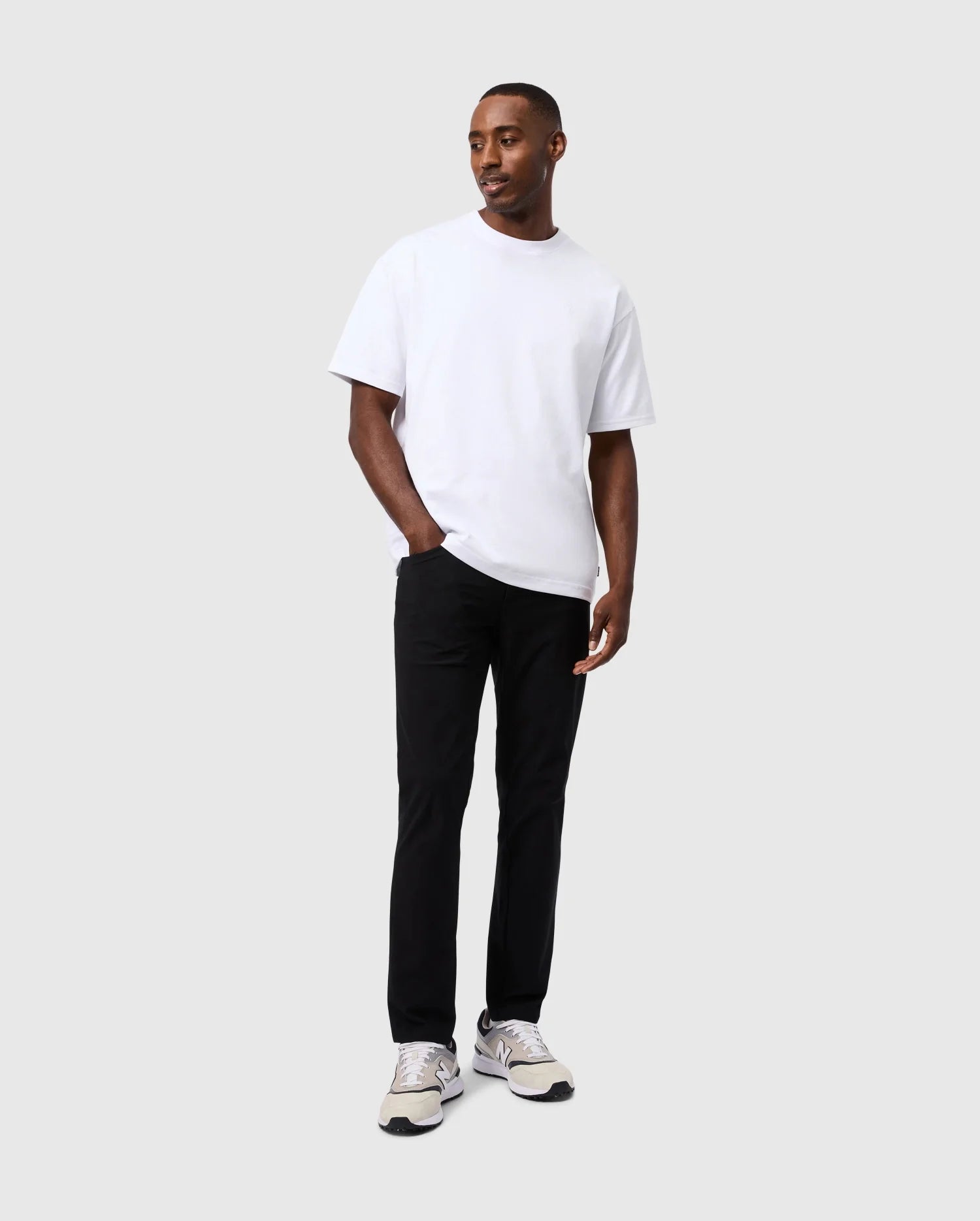 A person wearing a white t-shirt, the Psycho Bunny Mens Maverick 10K Pant - B6P788D200 with four-way stretch, and white sneakers stands against a plain gray background, looking slightly to the side with hands in pockets.