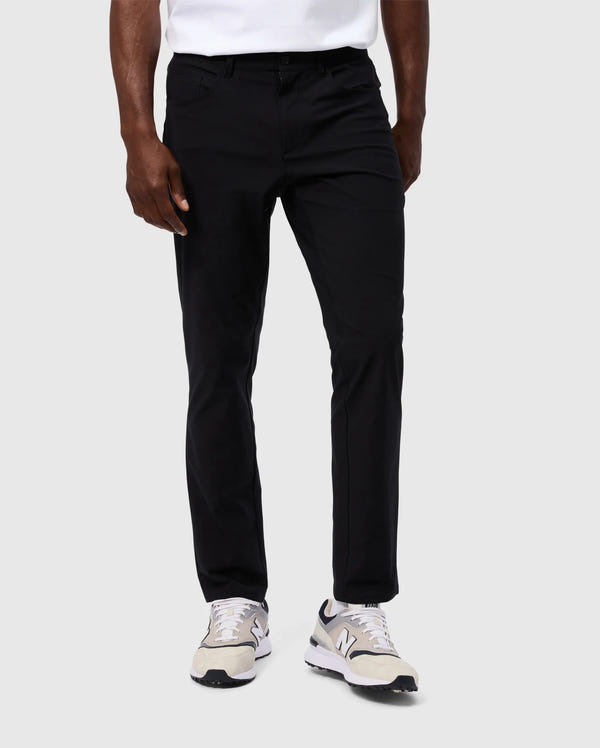 A person stands against a plain white background, wearing sleek black Psycho Bunny MENS MAVERICK 10K PANT - B6P788D200 and white sneakers with gray accents. The upper part of the person is not visible.