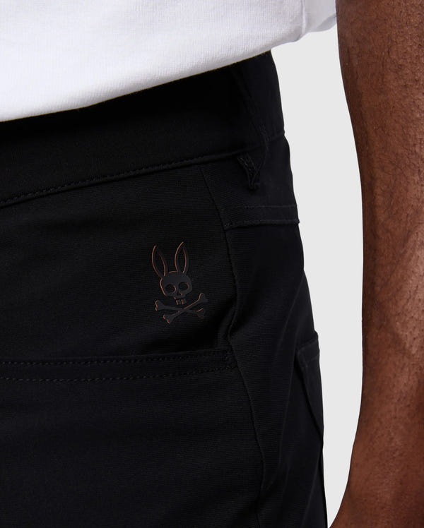 A close-up view showcases someone wearing Psycho Bunny's MENS MAVERICK 10K PANT - B6P788D200 in black, highlighted by the distinctive skull and bunny ears logo on the pocket. These four-way stretch pants offer a blend of comfort and style, impeccably paired with their crisp white shirt.