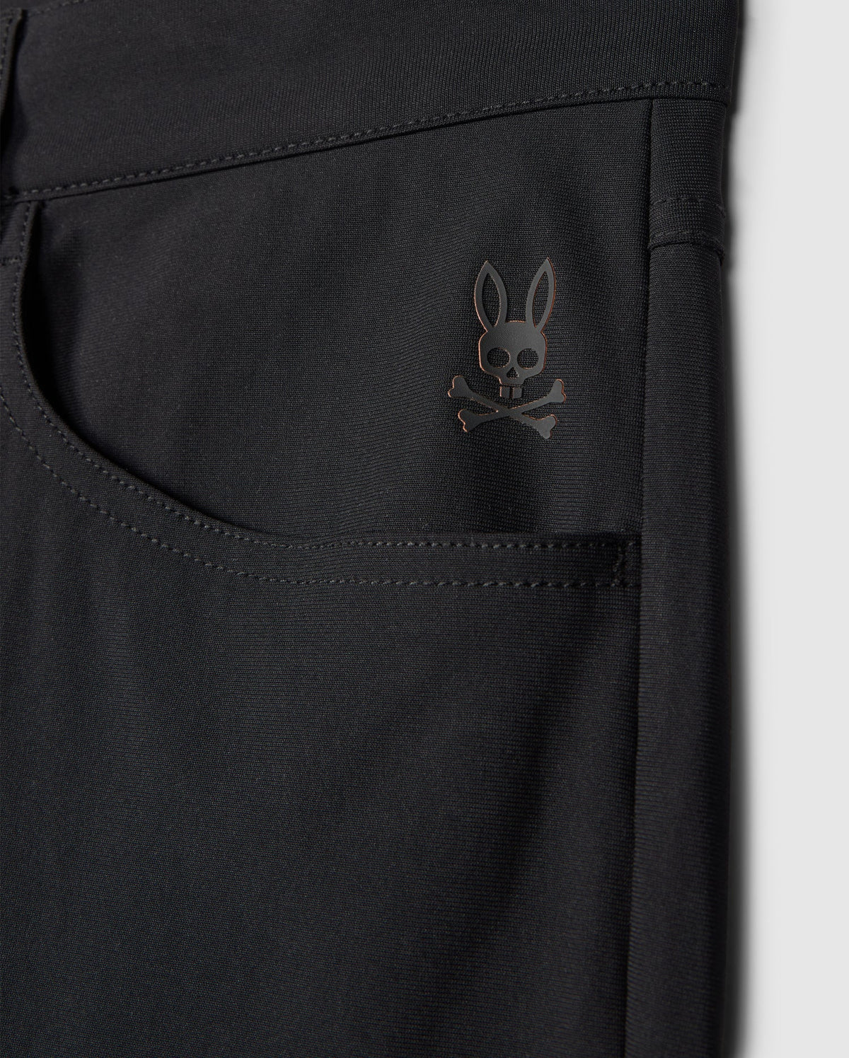 Close-up of the Psycho Bunny MENS MAVERICK 10K PANT - B6P788D200 in black highlighting a pocket adorned with a subtle skull and crossbones logo topped with bunny ears near the upper corner. The fabric looks smooth, slightly glossy, and incorporates four-way stretch for enhanced comfort.