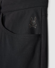 Close-up of the Psycho Bunny MENS MAVERICK 10K PANT - B6P788D200 in black highlighting a pocket adorned with a subtle skull and crossbones logo topped with bunny ears near the upper corner. The fabric looks smooth, slightly glossy, and incorporates four-way stretch for enhanced comfort.