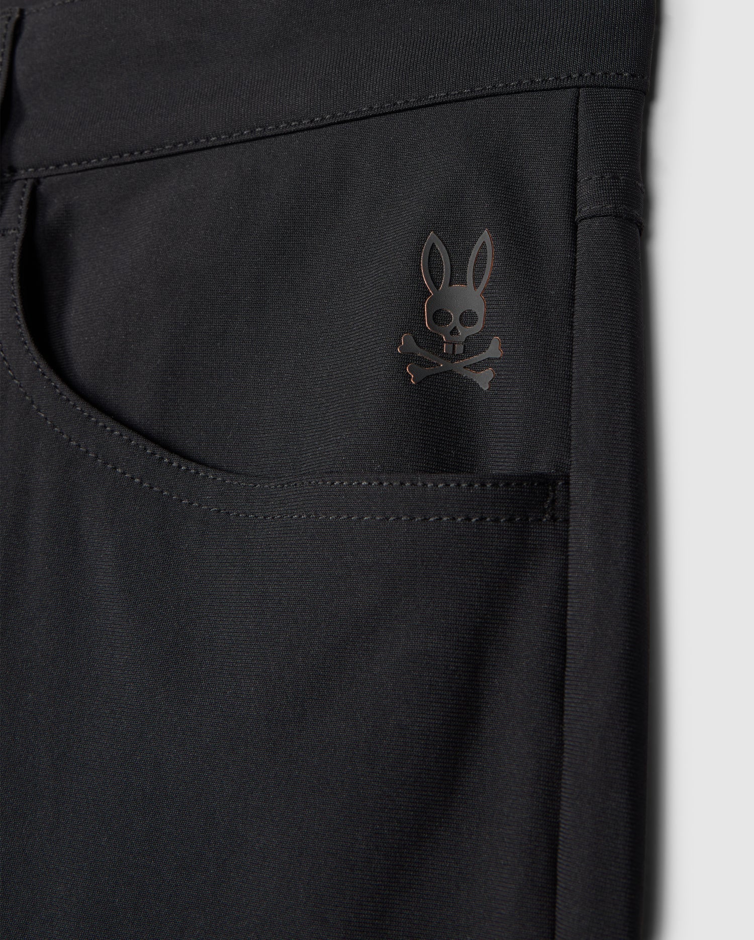 Close-up of the Psycho Bunny MENS MAVERICK 10K PANT - B6P788D200 in black highlighting a pocket adorned with a subtle skull and crossbones logo topped with bunny ears near the upper corner. The fabric looks smooth, slightly glossy, and incorporates four-way stretch for enhanced comfort.