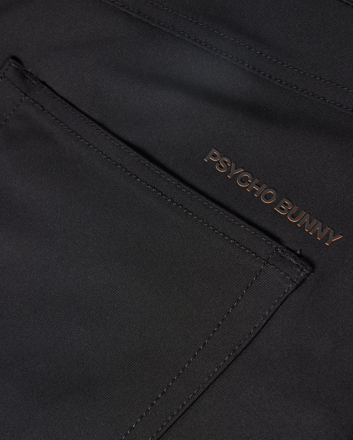 The MENS MAVERICK 10K PANT - B6P788D200 from Psycho Bunny showcases a sleek pocket in a close-up of the dark fabric. The brand name "PSYCHO BUNNY" is embroidered in a contrasting color near the edge, enhancing its modern design with four-way stretch for ultimate comfort.