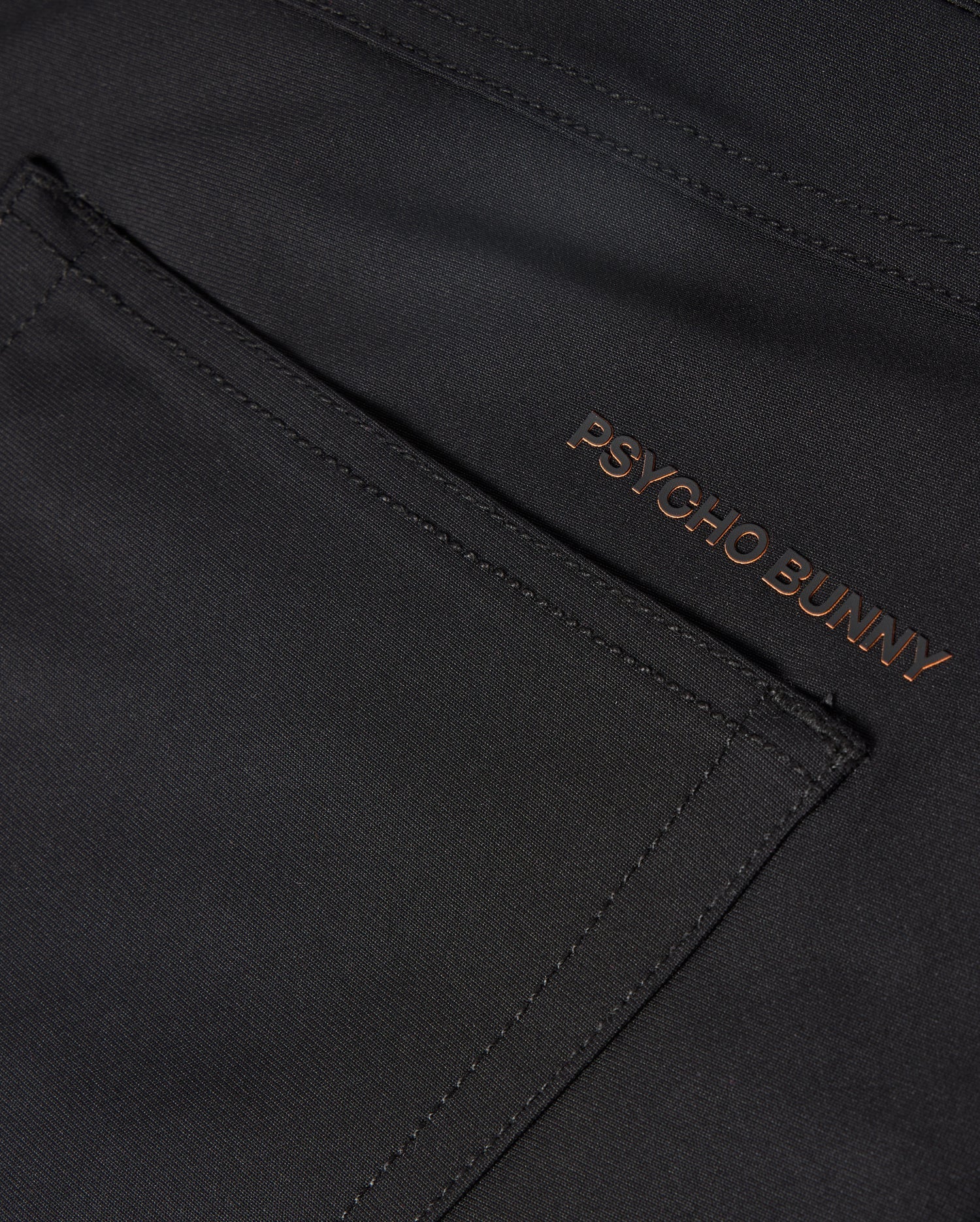 The MENS MAVERICK 10K PANT - B6P788D200 from Psycho Bunny showcases a sleek pocket in a close-up of the dark fabric. The brand name "PSYCHO BUNNY" is embroidered in a contrasting color near the edge, enhancing its modern design with four-way stretch for ultimate comfort.