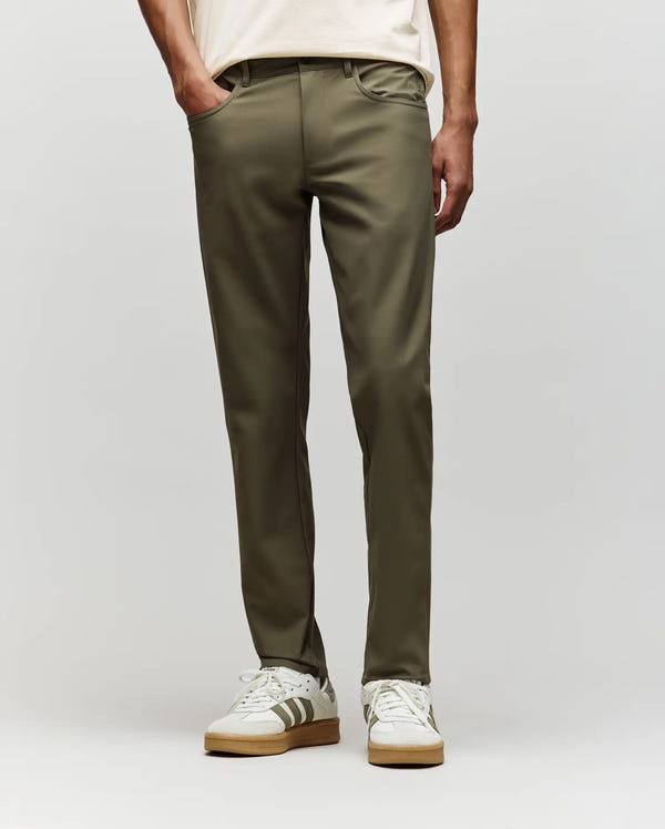 A person stands hands-in-pockets against a plain light background, wearing Psycho Bunny MENS MAVERICK 10K STEP PANTS - B6P788D200 in olive green, paired with a cream shirt and white sneakers with gray stripes.