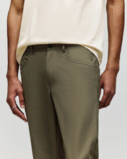 A person wears Psycho Bunny MENS MAVERICK 10K STEP PANTS - B6P788D200 in olive green with classic five-pocket styling and a cream shirt, standing with arms relaxed at their sides; only torso and upper legs are visible.