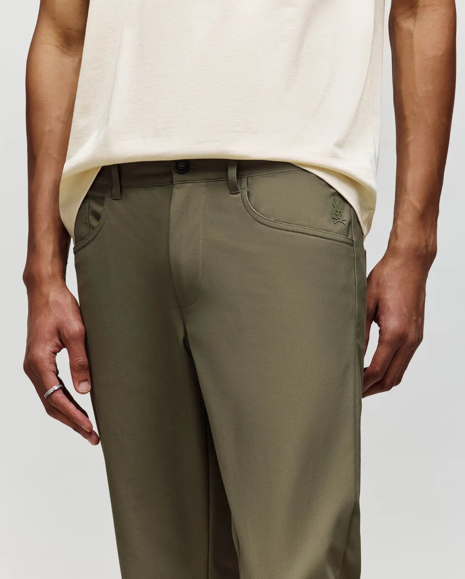 A person wears Psycho Bunny MENS MAVERICK 10K STEP PANTS - B6P788D200 in olive green with classic five-pocket styling and a cream shirt, standing with arms relaxed at their sides; only torso and upper legs are visible.