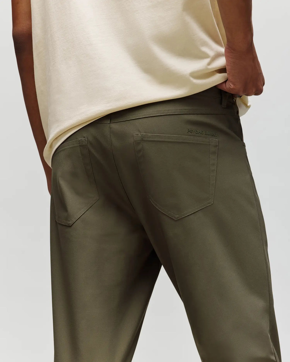 A person is seen from the back wearing a cream T-shirt and Psycho Bunny MENS MAVERICK 10K STEP PANTS - B6P788D200 in olive green, one hand in their pocket showcasing the pants' five-pocket design.