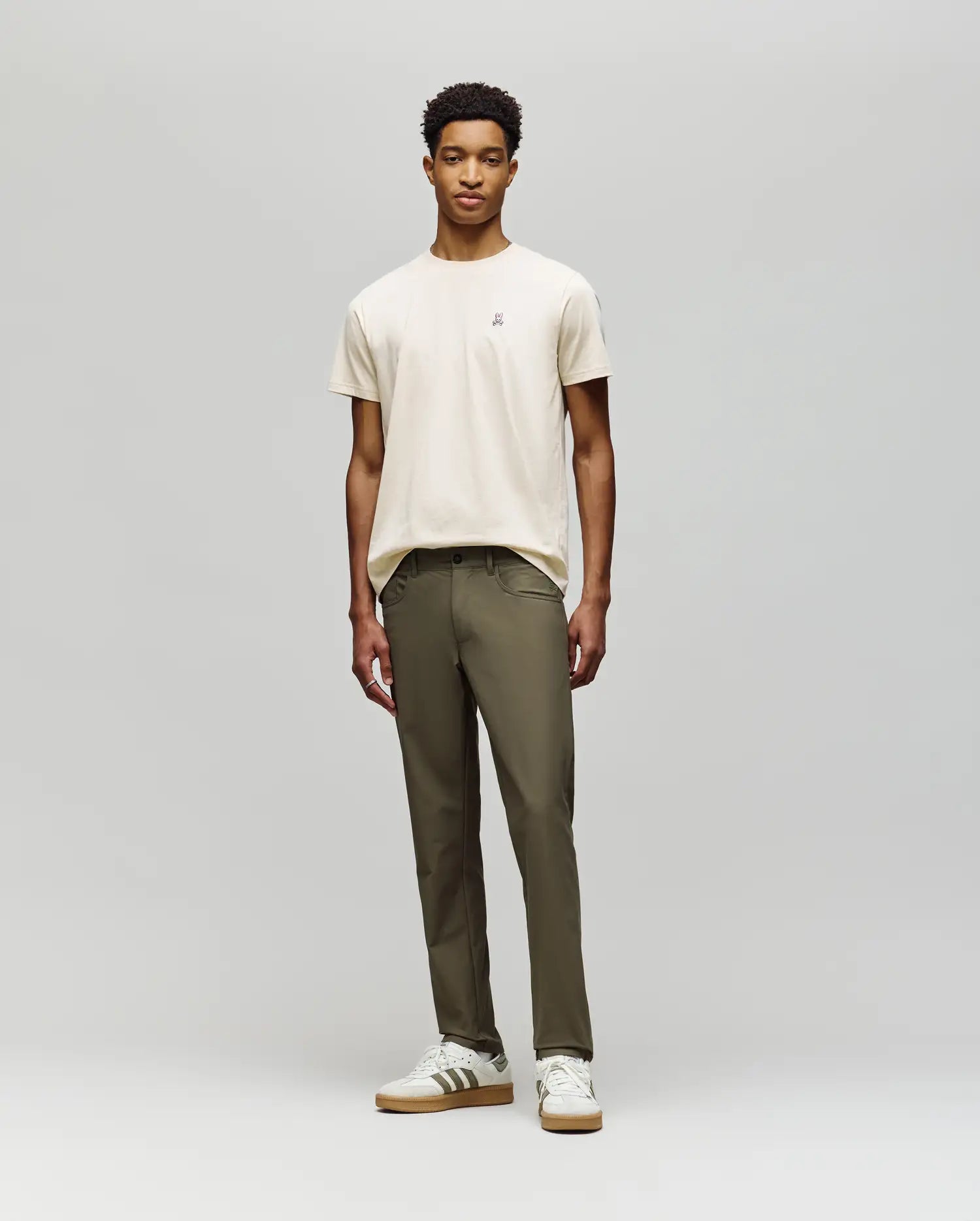A young man with short curly hair and a neutral expression stands against a light gray background, wearing a beige t-shirt, white sneakers with brown accents, and Psycho Bunny MENS MAVERICK 10K STEP PANTS - B6P788D200.