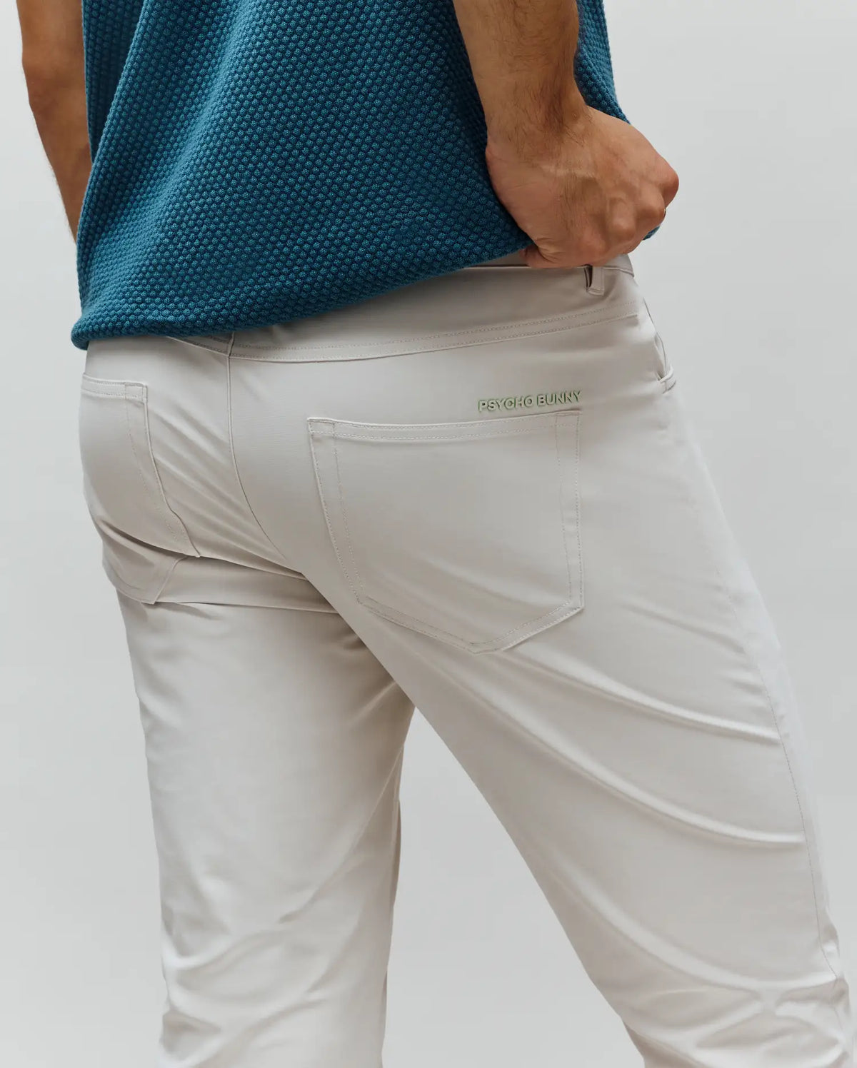 Mens Maverick 10k Step Pant in light beige, featuring a slim fit and smooth performance fabric. A small, light green embroidered PSYCHO BUNNY text logo is visible above the right back pocket.