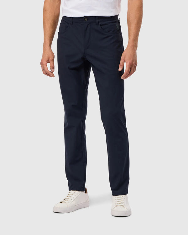 A person wearing dark blue MENS MAVERICK 10K PANT - B6P788D200 by Psycho Bunny and white sneakers stands against a light gray background, with the pants featuring a single button, paired with a tucked-in white shirt.